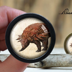 Pode incluir: Um close-up de um botão redondo de madeira preto com uma imagem detalhada de um dinossauro marrom e laranja com armadura espinhosa. O dinossauro está em um fundo creme com um design de estilo vintage.