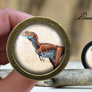 Pode incluir: Um par de botões redondos decorativos para armário com acabamento em bronze. Os botões apresentam uma imagem vintage de um dinossauro em um fundo creme.