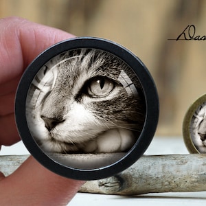 May include: Close-up of a black and white cat's face, looking to the right, in a round, black, framed, glass cabochon. The cabochon is on a wooden surface.