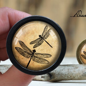 May include: Two black dragonfly silhouettes on a beige background with a vintage aesthetic. The image is encased in a black circular frame.