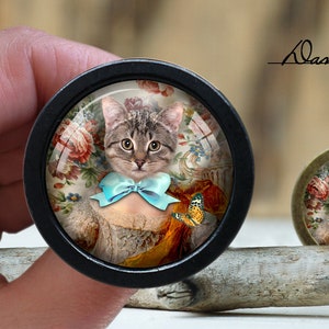 Peut inclure: Un cabochon rond en verre encadré de noir avec un chat tabby gris portant un nœud papillon bleu et un papillon. Le chat est entouré d'un motif floral.