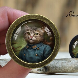 May include: A gold-toned round pendant with a glass dome featuring a portrait of a cat wearing a blue uniform. The cat has brown and white fur and is looking directly at the viewer. The pendant is on a wooden surface. "Dan's diary" is written in black script above the pendant.