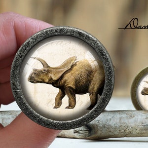 Pode incluir: Um close-up de um botão redondo, prateado com uma imagem vintage de um triceratops marrom. O botão está em uma superfície de madeira.