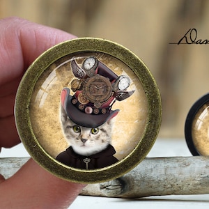 May include: A round, gold-coloured, framed glass cabochon featuring a steampunk-style cat wearing a top hat adorned with gears and clock faces. The cat is looking at the viewer with a curious expression. The background is a vintage-style parchment paper.