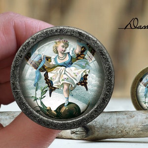 May include: Two round, antique-style cabochons with a vintage illustration of a woman with butterfly wings. The woman is wearing a blue and white dress. The cabochons have a dark, textured metal frame. The text "Dan's diary" is written in cursive.