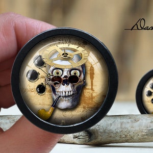May include: A pair of black ear gauges featuring a skull design. The skull has a clockwork design on its head and is wearing glasses. The gauges are displayed on a wooden surface.