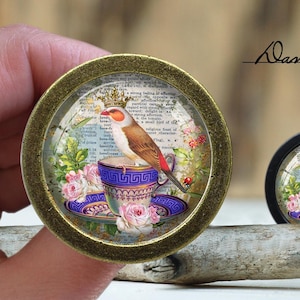 May include: A gold-toned round pendant with a glass dome featuring a bird perched on a teacup. The bird is brown and white with a red beak and a crown on its head. The teacup is blue and white with a floral pattern. The background is a vintage book page with text and floral illustrations.