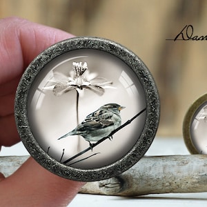 Pode incluir: Um pingente redondo prateado com uma cúpula de vidro que apresenta uma imagem em preto e branco de um pequeno pássaro empoleirado em um galho com uma delicada flor branca.