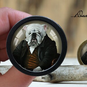 May include: A white bulldog wearing a black suit and a white cravat, with a serious expression, is depicted in a round glass cabochon. The cabochon is set in a black metal frame.