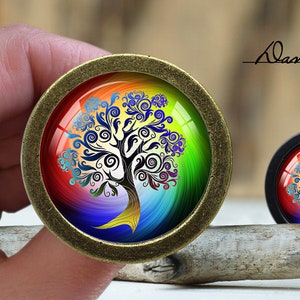 May include: A round, bronze-colored pendant with a colorful, rainbow-like background and a black tree of life design. The tree has intricate swirls and branches. The pendant is on a wooden surface.