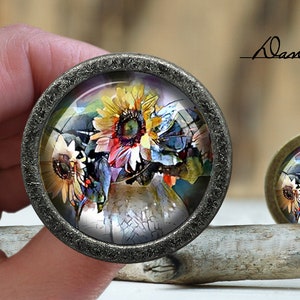 Puede incluir: Un colgante redondo de tono plateado con una cúpula de vidrio que presenta una pintura acuarela colorida de girasoles. El colgante está sobre una superficie de madera.