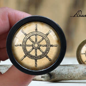 Puede incluir: Un pomo decorativo negro y dorado con un diseño de rueda náutica vintage. La rueda está detallada con radios y un cubo central.