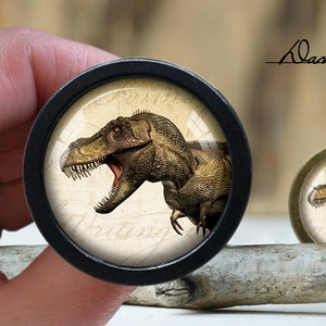 May include: A black and gold round button featuring a vintage-style image of a dinosaur with its mouth open. The dinosaur is brown and green and is set against a cream background with the word "Writing" visible.