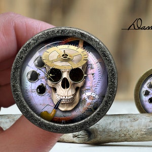 May include: A silver-toned round pendant featuring a skull design. The skull is wearing goggles and a clockwork gear on its head. The background is a purple and blue design with gears and clock hands.