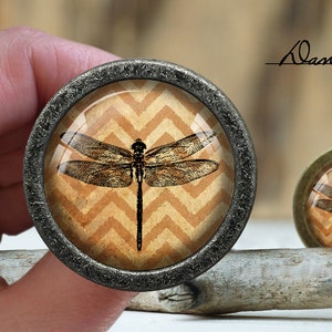 May include: A decorative cabinet knob with a vintage dragonfly design on a chevron patterned background. The knob is made of metal and has a dark gray finish.