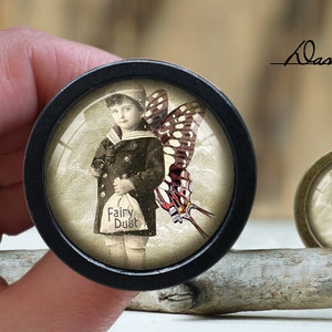 May include: A black and gold round knob with a glass insert featuring a vintage image of a child wearing a blue jacket and holding a bag that says "Fairy Dust". The child is standing in front of a large butterfly with red, white, and black wings.