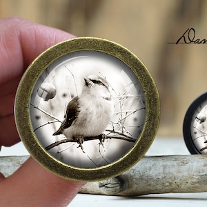 May include: A round, bronze-coloured pendant with a glass dome featuring a black and white photograph of a small bird perched on a branch in a snowy forest. The pendant is on a wooden surface.
