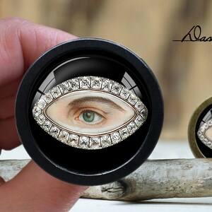 May include: A black circular box with a glass lid containing a detailed eye with a diamond-encrusted frame. The eye is painted with a realistic style and features a green iris. The box is sitting on a wooden surface.