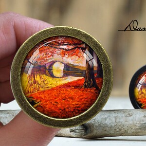 May include: Two round, decorative cabinet knobs with a landscape painting under a glass dome. The painting features a path through a forest with red and yellow autumn leaves. The knobs have bronze and black metal frames. The text "Dan's diary" is written in cursive.