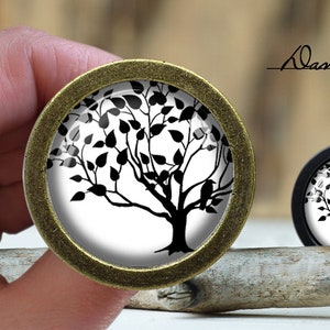 May include: A black and white silhouette of a tree with leaves and birds on a white background. The image is on a round, gold-coloured metal button.
