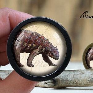 Op de afbeelding: Een zwarte en zilveren knop met een ronde, transparante glazen inzet. De glazen inzet toont een gedetailleerd beeld van een rode en bruine dinosaurus met stekels op zijn rug.