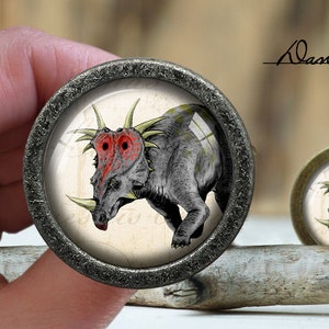 Pode incluir: Close-up de um botão redondo decorativo de cor prateada com um inserto de vidro que apresenta uma ilustração detalhada de um dinossauro cinza com chifres vermelhos e espinhos amarelos. O dinossauro está sobre um fundo branco com texto preto.