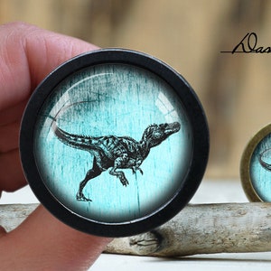 Pode incluir: Um botão preto com um fundo azul turquesa que apresenta uma silhueta preta de um dinossauro. O dinossauro está olhando para a direita e tem uma cauda longa. O botão está sobre uma superfície de madeira.