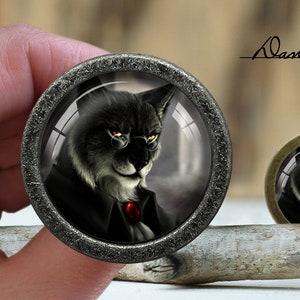 Peut inclure: Un bouton décoratif rond, de couleur argentée, recouvert de verre, représentant un chat noir avec un nœud papillon rouge et des lunettes. Le bouton est posé sur une surface en bois.