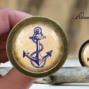 May include: A close-up of a gold-toned round knob with a glass insert featuring a blue anchor illustration on a beige background. The knob is on a wooden surface.