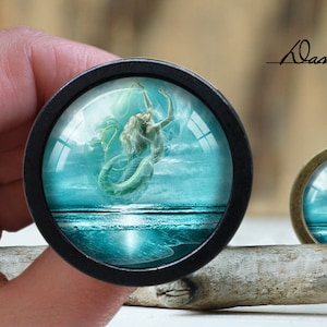 May include: A black and silver round button with a glass dome featuring a mermaid with a blue tail swimming in a turquoise ocean with white clouds. The button is on a wooden surface.