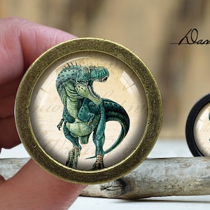 以下が含まれることがあります： 緑色の恐竜のイラストが描かれた、金色の丸いノブ。恐竜は右を向いており、白い背景に黒い文字が書かれています。