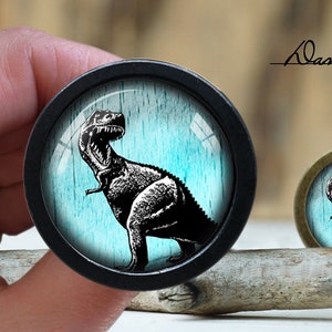 Pode incluir: Dois cabochons redondos, pretos e bronze, com tema de dinossauro. O cabochon maior é segurado por uma mão. Cada cabochon apresenta uma ilustração em preto e branco de um Tyrannosaurus Rex contra um fundo azul. O texto "Alan's diary" é visível.