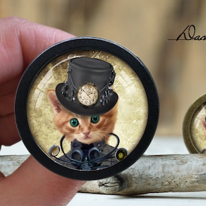 Op de afbeelding: Een close-up van een ronde, zwart geframed, glazen cabochon met een steampunk-achtige oranje tabby kat die een zwarte hoge hoed draagt met een klok gezicht en een bril. De kat kijkt recht naar de kijker. De achtergrond is een vintage-stijl, crèmekleurig papier met een vervaagd, cirkelvormig patroon.