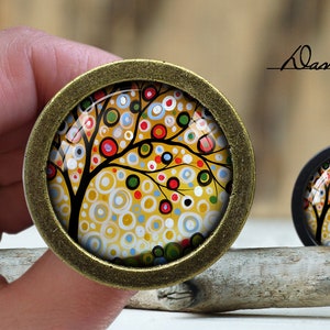 Könnte beinhalten: Ein goldfarbener Metallanhänger mit einem farbenfrohen abstrakten Baumdesign. Der Baum hat einen braunen Stamm und Äste mit weißen, gelben, roten und grünen Kreisen. Der Anhänger liegt auf einer Holzoberfläche.