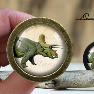 Pode incluir: Um botão de latão com um inserto redondo de vidro transparente que apresenta uma ilustração vintage de um dinossauro Triceratops verde e amarelo. O dinossauro é retratado em um fundo creme com a palavra "Mammoth" escrita abaixo.