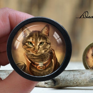 Op de afbeelding: Een close-up van een ronde, zwart geframed glazen cabochon met een afbeelding van een tabbykat die een leren halsband draagt met een metalen gesp. De kat kijkt recht in de camera met een serieuze uitdrukking. De achtergrond is wazig, lichtbruin van kleur. De cabochon ligt op een stuk drijfhout.