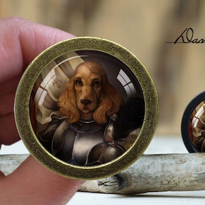 May include: A round, gold-colored, decorative pin with a glass dome featuring a painting of a brown and white dog wearing a suit of armor. The dog is looking directly at the viewer. The pin is on a wooden surface.