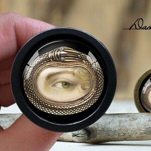 Könnte beinhalten: Ein runder Knopf aus schwarzem Holz mit einem goldenen und schwarzen Schlangendesign. Die Schlange ist um ein Kamee mit einem sichtbaren Frauenauge gewickelt. Der Knopf befindet sich auf einer Holzoberfläche.