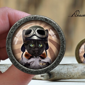 Peut inclure: Un chat noir portant un casque d'aviateur en cuir marron et des lunettes, avec un col en fausse fourrure marron et une veste de vol grise, est représenté sur un fond rayé marron et blanc. L'image est encadrée dans un cercle en métal argenté.