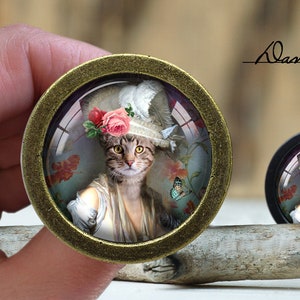 May include: A round, gold-toned, framed glass cabochon featuring a portrait of a cat wearing a hat adorned with pink flowers and a white feather. The cat is looking directly at the viewer. The background is a floral pattern.