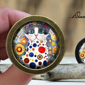 May include: A black and white cat with colorful polka dots on a white background. The cat is surrounded by colorful circles and swirls. The image is framed in a gold metal circle.