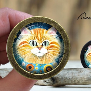 May include: A round, gold-toned metal pin with a glass cabochon featuring a painting of a fluffy orange cat with green eyes. The cat is surrounded by a blue and white background with colorful dots and swirls. The pin is on a wooden surface.