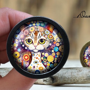 May include: Two decorative cat-themed cabochons. The larger one is held by a hand. The cat has a colourful, abstract design with yellow eyes and a white chest. The smaller cabochon is resting on a piece of wood. The text "Oasi's diary" is visible.