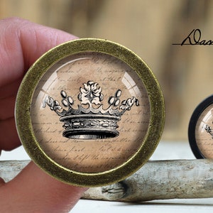 Può includere: Due pomelli decorativi per mobili con un design a corona vintage. Uno ha un bordo in metallo color bronzo, l'altro un bordo nero. La corona è un'illustrazione dettagliata in bianco e nero su sfondo beige con scritte.