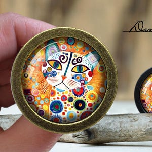 May include: Two decorative cabinet knobs featuring a colourful cat face design. The knobs have a bronze finish and a glass cabochon with a vibrant cat illustration. The cat's face is white with blue, yellow, and orange accents. The text "Dan's diary" is visible.