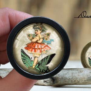 May include: A black framed round glass cabochon with a vintage illustration of a fairy with wings and a red dress. The fairy is holding a wand and standing on a green butterfly. The background is a light beige color.