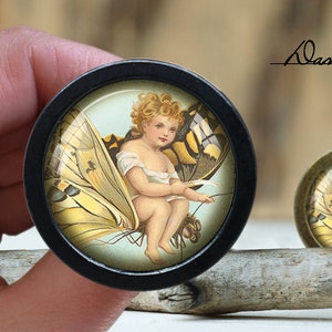 May include: A black wooden round button with a glass insert featuring a young girl with wings, sitting on a butterfly with yellow and brown wings. The girl is wearing a white dress and has blonde hair.