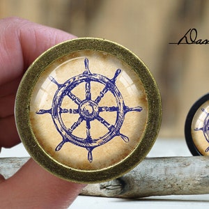 May include: A gold-toned round knob with a glass insert featuring a blue and white nautical wheel design. The wheel is set against a beige background.