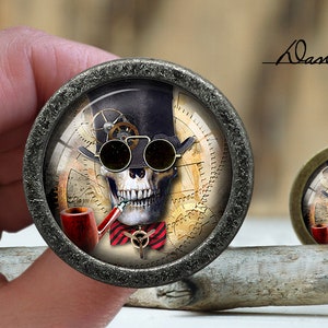 May include: A round, silver-toned metal button featuring a steampunk skull design. The skull is wearing a top hat and goggles, and is holding a pipe. The background is a clock face with gears and cogs.