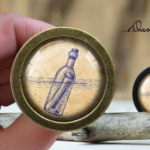 May include: A brass knob with a glass insert featuring a blue and white illustration of a message in a bottle floating in the water. The text "Dan's diary" is visible in the background.